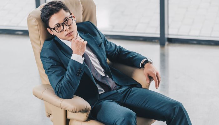 Confident asian businessman in eyeglasses sitting with hand on chin in office